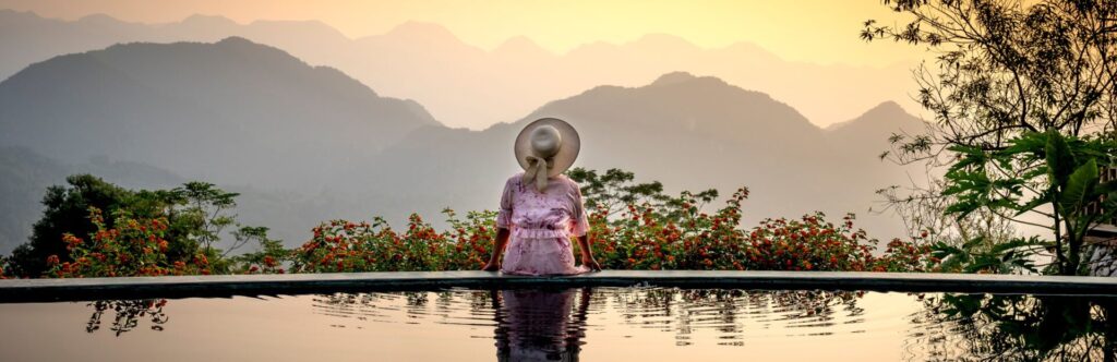 Persona de espaldas sentada en ele borde de una piscina con un sombrero y mirando a las montañas y atardecer