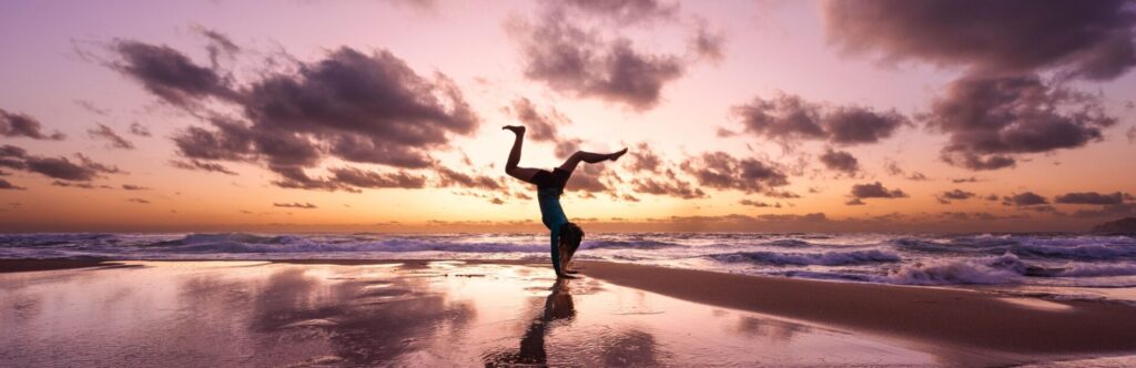 Persona en la playa a la orilla del mar haciendo el pino con el atardecer de fondo